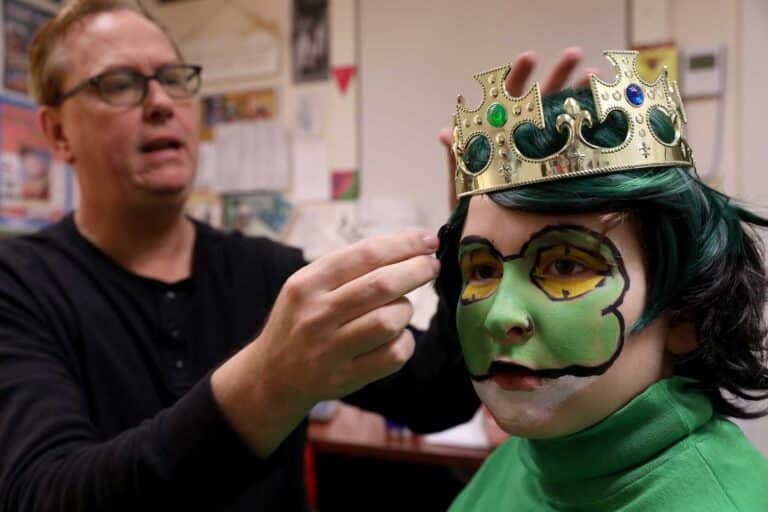 A teacher applies stage makeup to a student wearing a crown